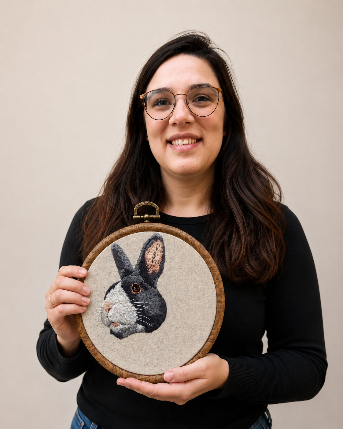 Bianca Kau in her studio, holding a hand-embroidered hoop portrait of a black-and-white rabbit on natural linen.