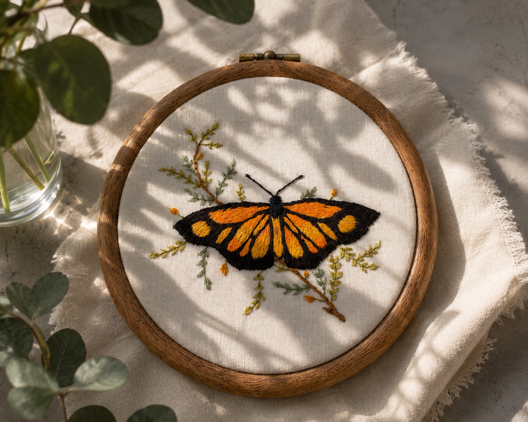 A monarch butterfly hand-embroidered in orange and black, framed by fine botanical sprigs on linen.
