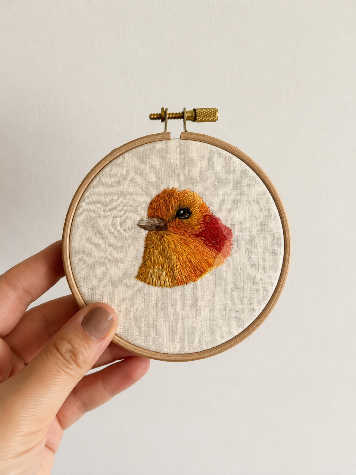 A small finch hand-embroidered in golden-orange thread with a red cheek patch and grey beak, in a pale wooden hoop.