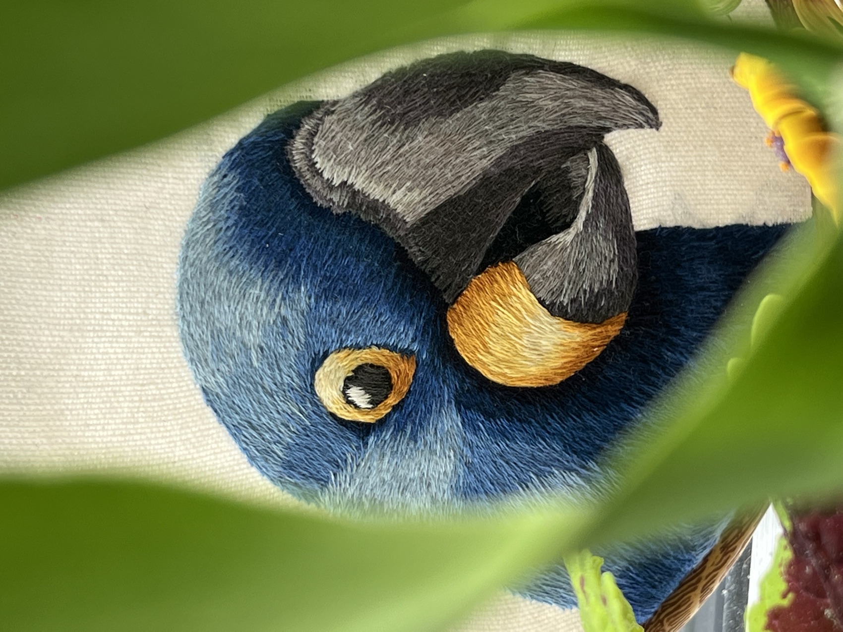A hyacinth macaw, hand-embroidered in deep cobalt-blue thread with a grey beak and golden eye-ring, glimpsed through soft green leaves.