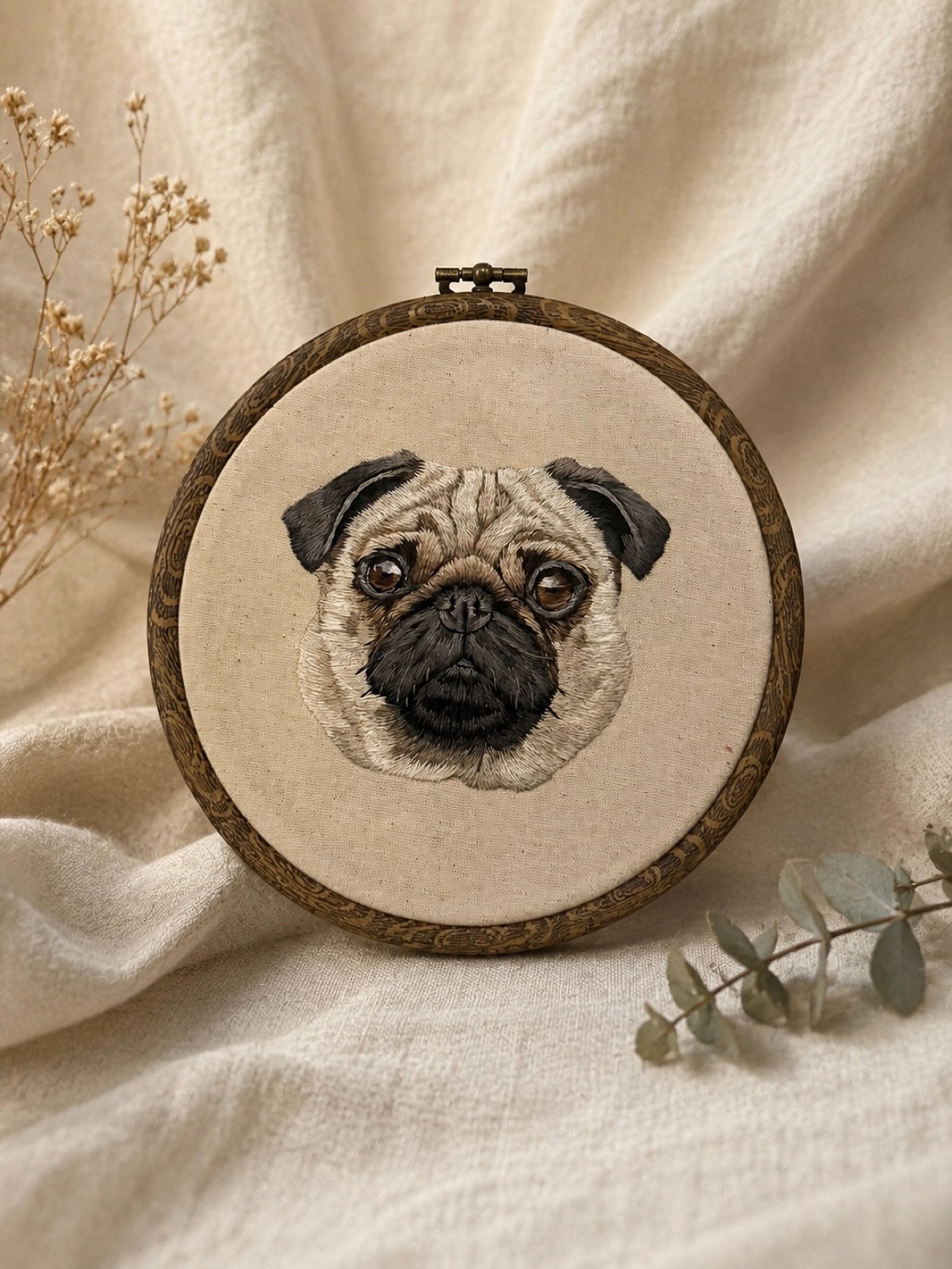 A pug hand-embroidered in soft fawn and charcoal thread on raw cotton, set in a dark wooden hoop.